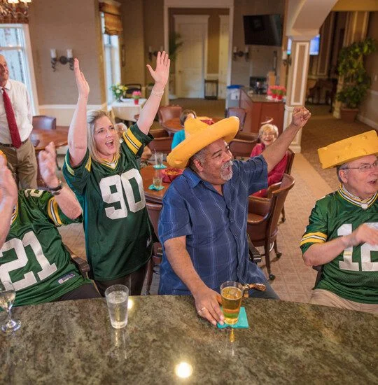 smiling group of seniors, friends, and team members cheer on the Green Bay Packers while watching a game in the lounge