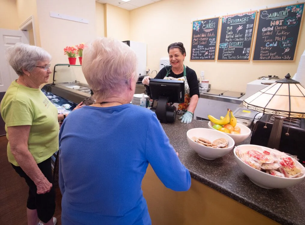 two smiling senior woman place an order at the Castle Cafe dining venue in Newcastle Place, served by a cheerful team member