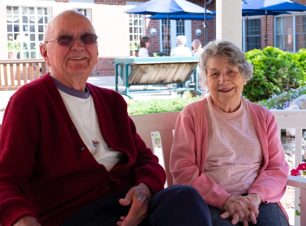 smiling senior couple sits on a bench outdoors at Newcastle Place Senior Living Community