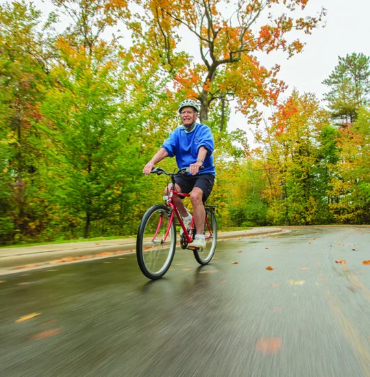 smiling senior man in athletic gear and helmet rides bicycle along a tree-lined road in autumn