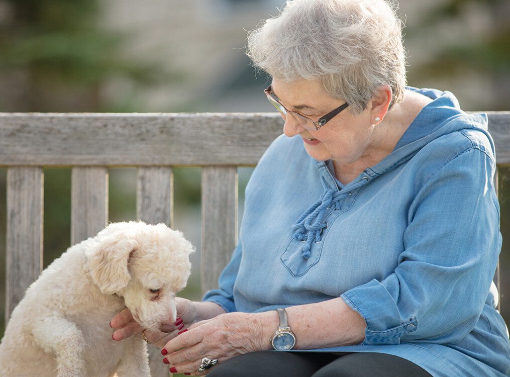 happy senior woman in blue shirt pets her small dog while seated on an outdoor bench at Newcastle Place