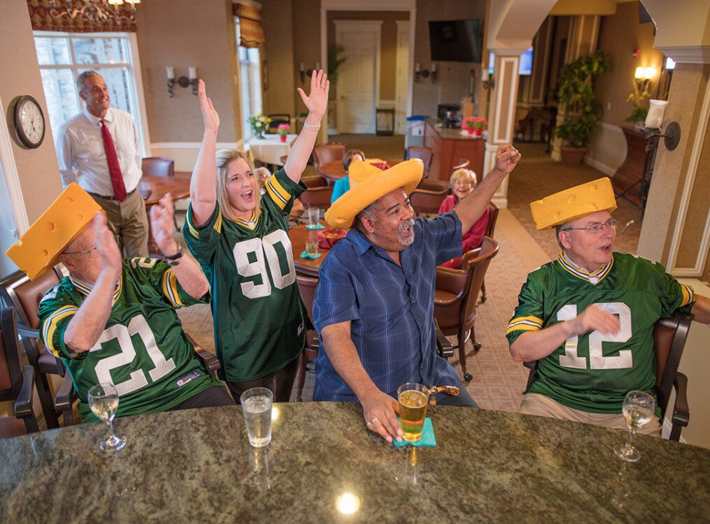 smiling group of seniors, friends, and team members cheer on the Green Bay Packers while watching a game in the lounge