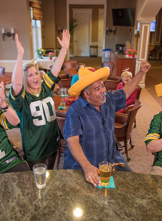 smiling group of seniors, friends, and team members cheer on the Green Bay Packers while watching a game in the lounge