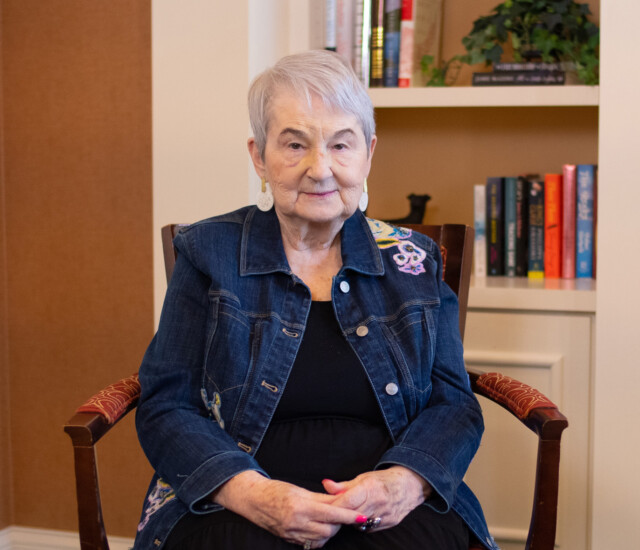 smiling senior woman, Gail Johnson, in denim jacket sits in front of a bookshelf for an interview at Newcastle Place