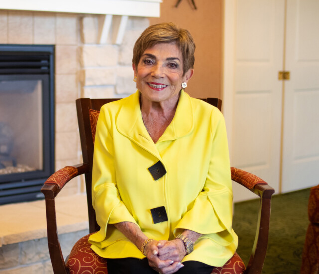 smiling senior woman, Marlene Lauwasser, in yellow blouse sits in front of a fireplace for an interview at Newcastle Place
