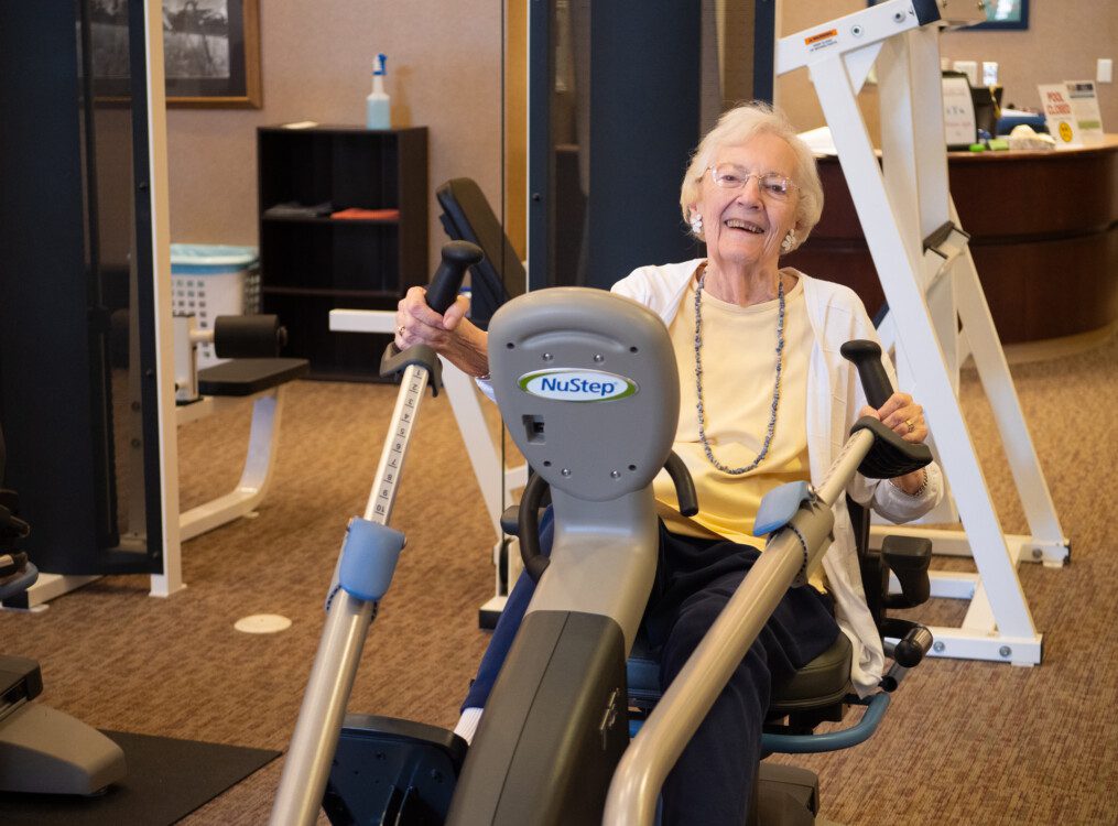 smiling senior woman in casual clothing uses a seated elliptical machine in the gym at Newcastle Place Senior Living Community