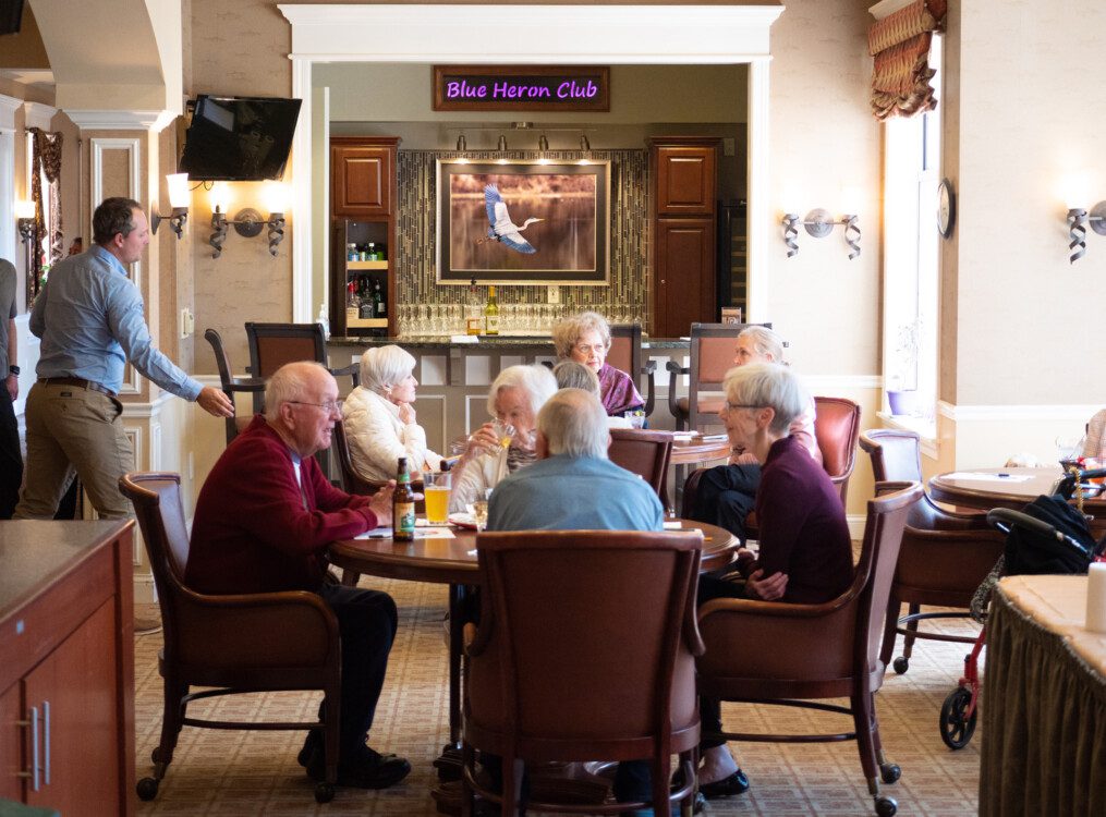 Two tables of happy senior friends dine in at the elegant Blue Heron Club venue and lounge at Newcastle Place Senior Living Community