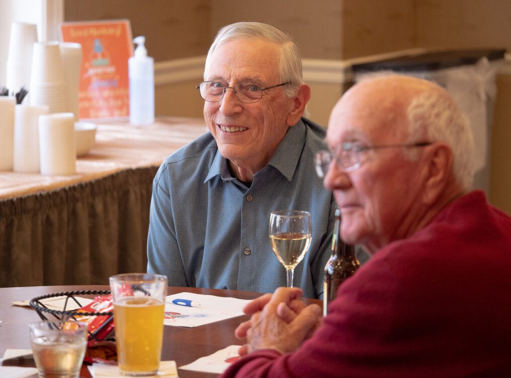 two smiling senior men enjoy wine and a meal with friends at a dining venue in Newcastle Place Senior Living Community
