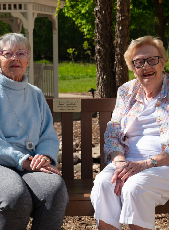 two smiling senior women sit on a memorial bench in front of a gazebo at Newcastle Place Senior Living Community