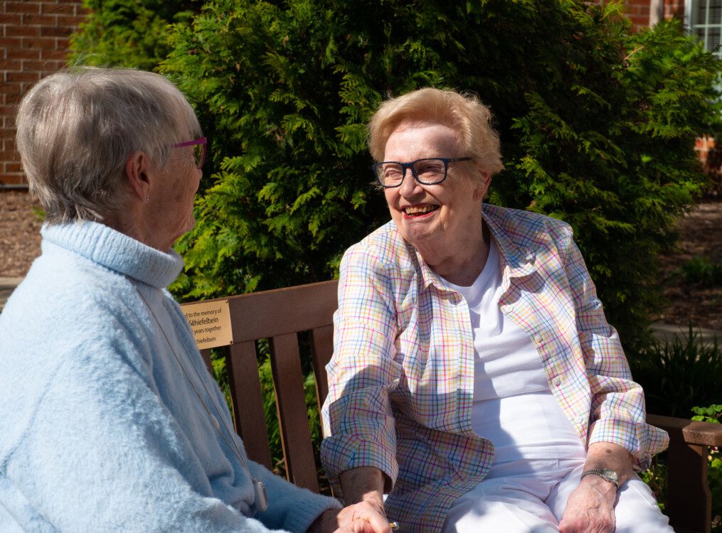two cheerful senior women converse while seated on a memorial bench outside in the sunshine at Newcastle Place Senior Living Community