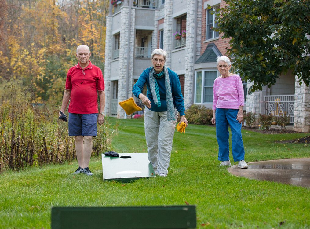 group of senior friends smile and play a game of cornhole outdoors in front of their apartments at Newcastle Place Senior Living Community